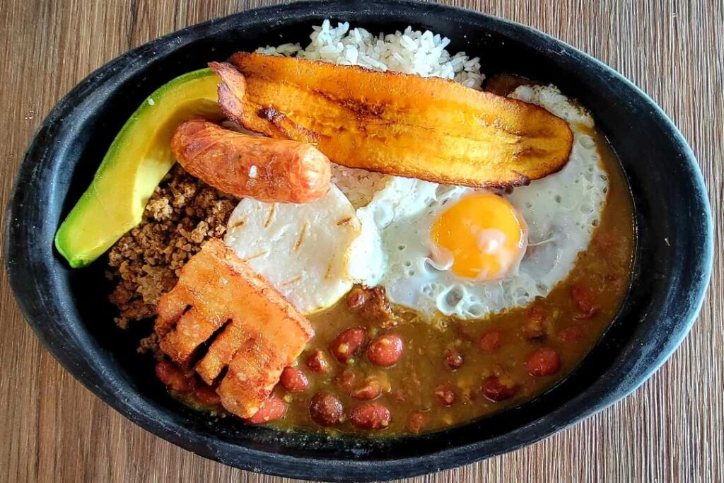 Colombian sausages, ground beef, rice, red beans, crispy fried pork, egg and avocado in a black plate